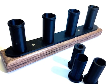 May include: A wooden and black shot glass holder with four upright cylindrical slots. The base is a rectangular piece of wood with a dark grain pattern. Four additional shot glass holders are placed near the base.