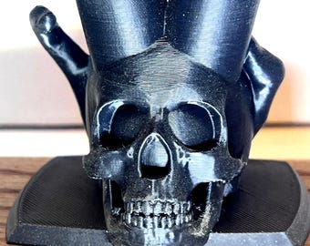May include: A black, 3D-printed skull sculpture with a detailed skeletal structure. The sculpture features a base and two upward-reaching elements, possibly for holding items. The skull's eye sockets and teeth are clearly defined.