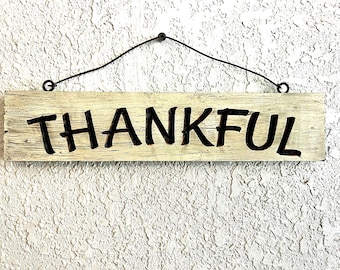May include: A rectangular wooden sign with the word "THANKFUL" in black, bold letters. The sign is cream-colored with a distressed finish and is suspended by a black wire. The background is a textured white wall.