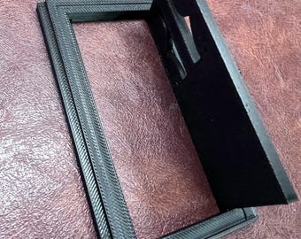 May include: A black rectangular picture frame with a textured border and a hinged back panel. The frame is open, revealing the interior space. The frame is made of a dark material and sits on a textured surface.