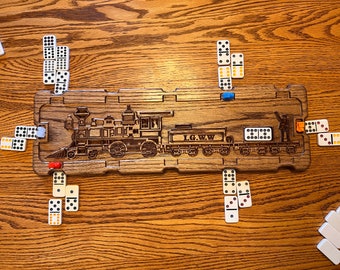 May include: A rectangular wooden Mexican Train dominoes hub featuring a laser-engraved steam locomotive and the text I.G.W.W. The board has notched edges for placing dominoes and includes small colored plastic markers positioned around the perimeter.