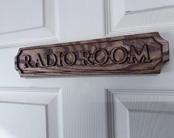 May include: A wooden sign with the words "RADIO ROOM" carved into it. The sign is rectangular with decorative edges and is mounted on a white door. The wood has a natural grain and a dark brown stain.