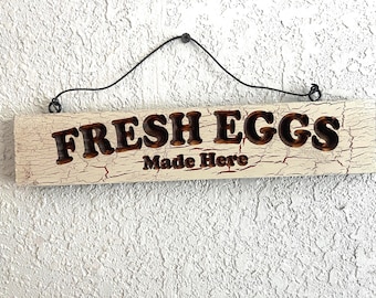 May include: A rectangular wooden sign with the words "FRESH EGGS Made Here" in dark brown lettering. The sign has a distressed, crackled cream-colored background and a black wire for hanging. The sign is mounted on a textured white wall.