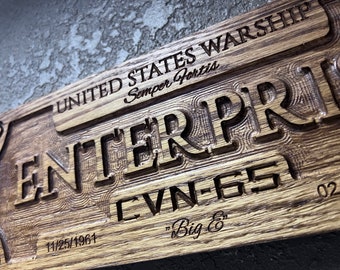 May include: A wooden plaque with carved text. The words "UNITED STATES WARSHIP" and "ENTERPRISE" are prominently displayed. Below, "CVN-65" and "Big E" are also carved. Dates "11/25/1961" and "02/03/2017" are included.