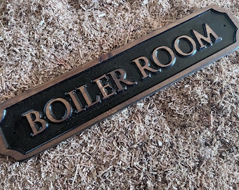 May include: A rectangular wooden sign with the words "BOILER ROOM" carved in large, raised letters. The sign has a black background with brown lettering and a brown border. The sign is resting on a bed of wood shavings.