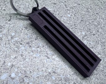 May include: A dark purple rectangular keychain with three parallel cut-out lines. The keychain is attached to a silver metal split ring. The item is resting on a speckled gray surface.