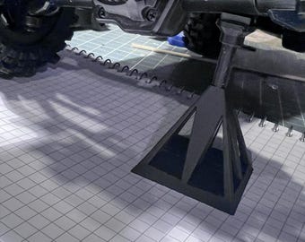 May include: A black, triangular jack stand supports the undercarriage of a black vehicle. The stand is on a white grid surface. The vehicle has a black chassis, a black axle, and a pink component.
