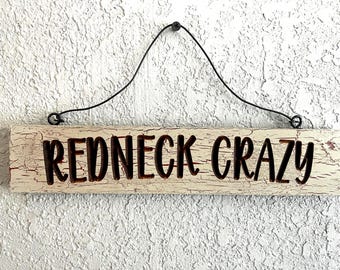 May include: A rectangular wooden sign with the words "REDNECK CRAZY" in dark brown lettering. The sign has a distressed, off-white finish with visible cracks. A black wire is attached for hanging.