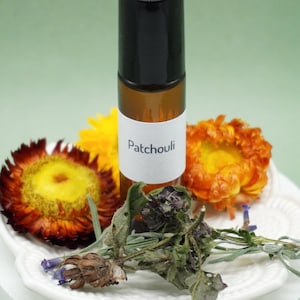 Puede incluir: Un frasco roll-on de vidrio marrón con tapa negra, etiquetado "Patchouli". El frasco está rodeado de flores secas en tonos amarillos, naranjas y rojos, y una ramita de hierbas secas. Los artículos están dispuestos sobre un plato blanco decorativo.