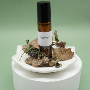 May include: A brown glass essential oil roller bottle with a black cap and a white label that reads "Vetiver." The bottle sits on a white, decorative plate surrounded by dried herbs and wood chips. The background is a solid green.
