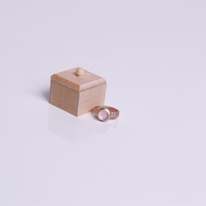 May include: A small wooden box with a lid and a light brown finish. The box is sitting on a white surface next to a gold-colored watch.