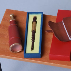 May include: A collection of miniature items: a red and gold telescope, a pen in a blue box, and a brown leather-bound book with a red stand. The objects are arranged on a wooden table, evoking a theme of exploration or study.