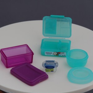 May include: A selection of small, colourful food storage containers. Includes a clear magenta rectangular box, a teal lunchbox with a hinged lid, a small clear container, and a teal bowl with a lid. The containers are on a white surface.