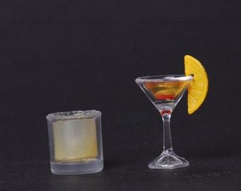 May include: A clear glass martini glass with a yellow citrus wedge garnish, filled with a light orange liquid, and a clear glass shot glass with a frosted rim on a black background.