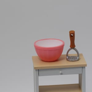 Könnte beinhalten: Eine Miniatur-Küchenszene mit einer rosa Schüssel und einem Kartoffelstampfer mit Holzgriff. Die Gegenstände stehen auf einem kleinen Holztisch mit weißem Rahmen und Schublade. Die Schüssel ist rund mit weißem Innenraum.
