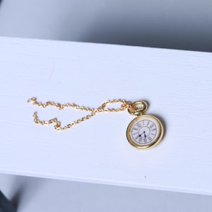 May include: A gold chain necklace with a small gold pocket watch pendant. The watch face has Roman numerals and black hands.