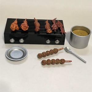 May include: Miniature black barbecue grill with six pieces of food, a small silver plate, a silver spoon, two skewers of food, and a silver container with food inside. The food on the grill is brown and orange.