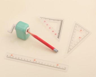 May include: A set of clear plastic school supplies including a ruler, two triangles, and a green pencil sharpener. A red pencil is lying on the white surface.