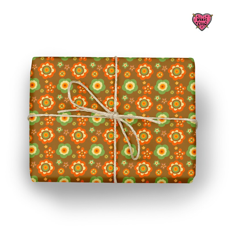 May include: A rectangular gift wrapped in brown paper featuring a repeating pattern of orange, light green, and yellow flowers. The package is tied with a thin, natural-colored jute twine string tied in a bow at the center. A small heart logo reads Pixie Drew.