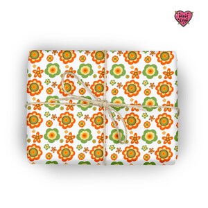 May include: A rectangular gift wrapped in white paper featuring a repeating pattern of orange, yellow, and green flowers. The package is tied with a thin, natural-colored jute twine string. A small heart-shaped logo with the text Pixie Drew is visible in the top right corner.