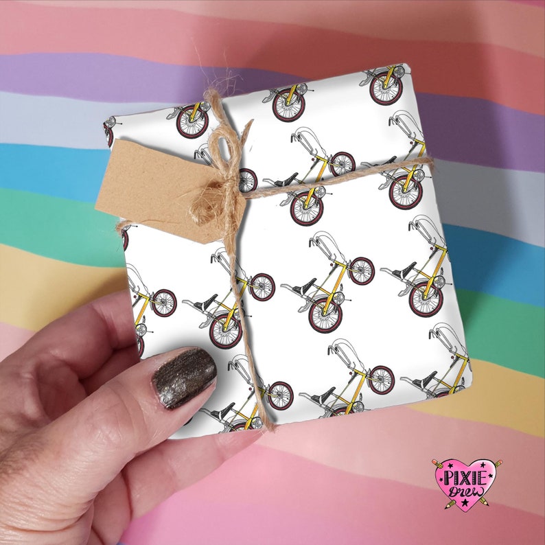 Op de afbeelding: Wit inpakpapier met een herhalend patroon van gele en zwarte fietsen. Het papier is om een ​​cadeaudoos gewikkeld en vastgebonden met touw. Een bruin cadeau-etiket is aan het touw bevestigd.