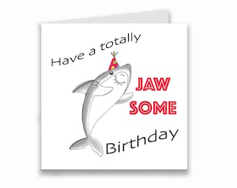May include: A white card with a gray shark wearing a red and yellow party hat. The text on the card reads "Have a totally jaw some birthday".