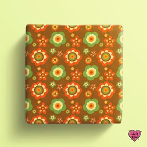 May include: A square gift box wrapped in brown paper featuring a repeating pattern of stylized flowers in shades of orange, light green, and cream. A small pink heart-shaped logo with the text Pixie Drew is visible in the bottom corner.