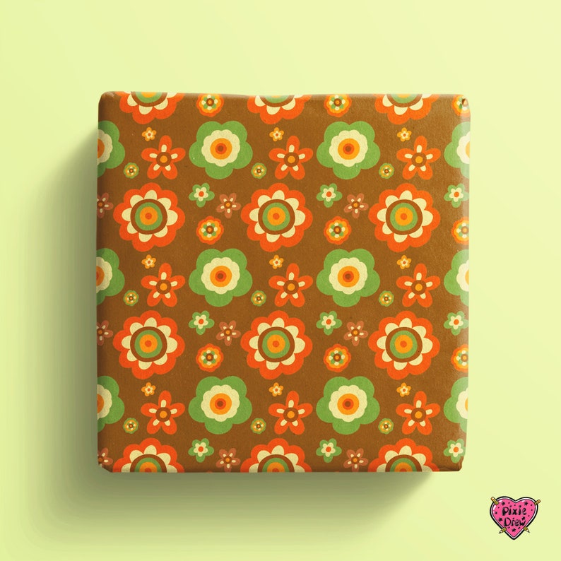May include: A square gift box wrapped in brown paper featuring a repeating pattern of stylized flowers in shades of orange, light green, and cream. A small pink heart-shaped logo with the text Pixie Drew is visible in the bottom corner.