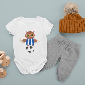 May include: A white baby onesie featuring a cartoon owl wearing a blue and white striped jersey and standing on a football. The onesie is paired with grey sweatpants and a brown knitted beanie with a fluffy pom-pom.