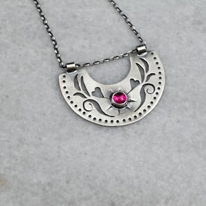 Può includere: Una collana color argento con un ciondolo a forma di semicerchio caratterizzato da motivi floreali e a cuore ritagliati. Il ciondolo è impreziosito da una singola pietra preziosa rotonda di colore rosa acceso al centro, appesa a una sottile catenina argentata.