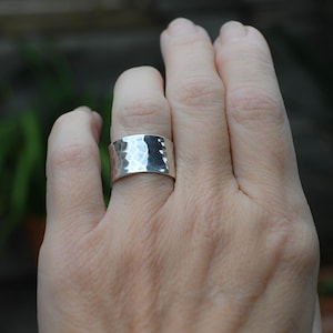 Könnte beinhalten: Ein breiter Silberring mit einer gehämmerten, texturierten Oberfläche, die ein reflektierendes, facettiertes Finish erzeugt. Das Metall hat einen polierten, metallischen Glanz und eine gleichmäßige Breite entlang des gesamten Ringumfangs.