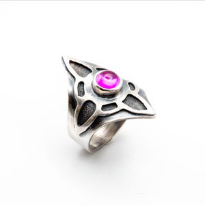 Pode incluir: Um anel de prata com um design triangular e uma pedra central redonda de cor magenta. O anel apresenta um padrão de nó celta e um acabamento texturizado, sobre um fundo branco.