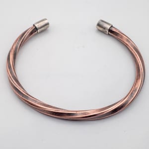 Peut inclure: Un bracelet manchette de couleur cuivrée avec un design torsadé. Le bracelet a une forme circulaire avec des embouts argentés à chaque extrémité. Le métal a un aspect texturé, et le style général est simple et élégant.