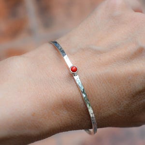 Può includere: Un braccialetto sottile color argento con una superficie martellata e testurizzata, caratterizzato da una piccola pietra rotonda rosso brillante incastonata in una lunetta circolare liscia al centro del cinturino, dal design essenziale.