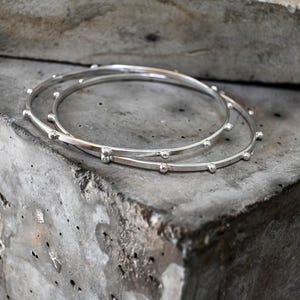 Może przedstawiać: Dwie cienkie, okrągłe srebrne bransoletki typu bangle z serią małych, równomiernie rozmieszczonych kulistych koralików wzdłuż zewnętrznej krawędzi każdej obręczy. Metalowa powierzchnia bransoletek ma polerowane wykończenie, które odbija światło.