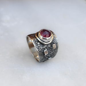 Può includere: Un anello a fascia larga con finitura in argento ossidato scuro, caratterizzato da una superficie testurizzata e craterizzata con piccoli accenti metallici. Una gemma rotonda, sfaccettata e rosso scuro è incastonata in una montatura liscia.