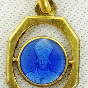 May include: Gold-toned pendant with an octagonal frame. Inside, a blue circular medallion features a portrait of a religious figure. The pendant has a loop for a chain. A religious themed jewelry item.
