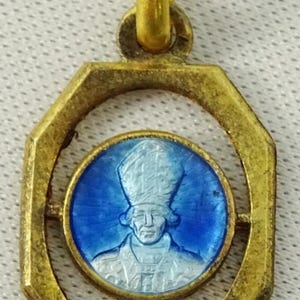 Peut inclure: Pendentif doré avec un cadre octogonal. À l'intérieur, un médaillon circulaire bleu présente une image détaillée d'une figure religieuse en blanc. Le pendentif a une boucle pour être suspendu.