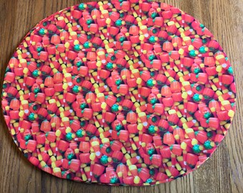 May include: A round orange, yellow, and green fabric placemat with a pattern of candy corn and pumpkin-shaped candies.