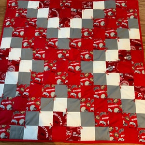 May include: A handmade patchwork quilt featuring a grid pattern of red, white, and gray squares. The red squares display a repeating pattern with the text "Buckeyes" and football helmets. The quilt has a red border.