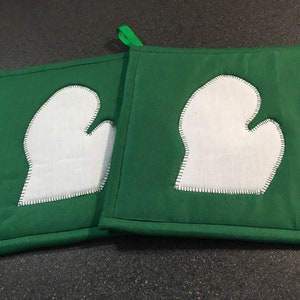 May include: Two green pot holders with white mitten shapes sewn on. The pot holders have a loop for hanging.