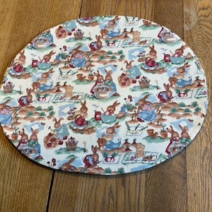 May include: A white oval placemat with a repeating pattern of bunnies in a garden setting. The bunnies are wearing blue and red clothing and are surrounded by flowers, plants, and vegetables.