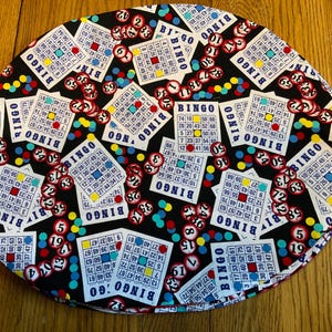 May include: Round black fabric with a repeating pattern of bingo cards, bingo chips, and colorful dots. The bingo cards are white with red, blue, yellow, and green accents. The bingo chips are red with white numbers.