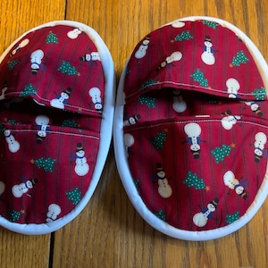 May include: A pair of red and white oven mitts with a snowman and Christmas tree pattern. The mitts have a pocket on the front.