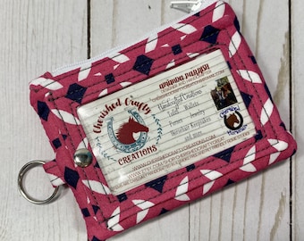 May include: A pink and white patterned cardholder with a clear ID window and a zip closure. The cardholder features a silver key ring and a snap closure. The cardholder has the text "Cherished Crafty Creations" and a horse logo.