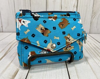 May include: A small, blue fabric wallet featuring a pattern of dogs, paw prints, and bones. It has a zip closure, a press stud, and a metal ring. The wallet is displayed on a metal stand. The wallet is approximately 10 cm wide.