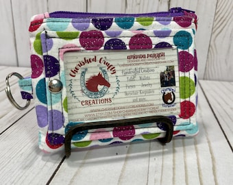 May include: A small, polka-dot wallet with a clear ID window. The wallet features a zip closure and a key ring attachment. The fabric has a white background with large, colourful polka dots in purple, pink, teal, and green. The wallet has text that reads "Cherished Crafty Creations."