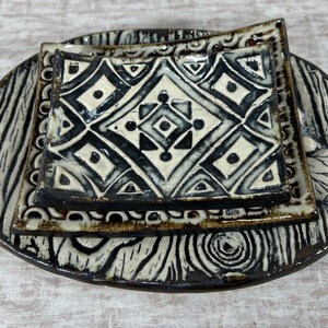 May include: A handcrafted ceramic serving set featuring a large oval platter with a square plate and a smaller square dish. The set has a black and white geometric pattern and a wood-grain texture. The set is ideal for serving appetizers or small dishes.