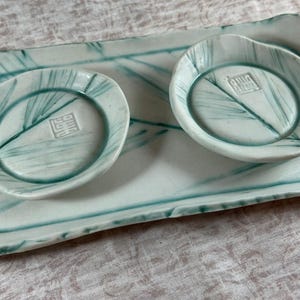 May include: A ceramic serving set featuring a rectangular tray and two small, round dishes. The set is white with teal-colored leaf-like patterns and a stamped symbol in each dish. The edges of the tray and dishes have a slightly irregular shape.