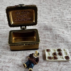 May include: A rectangular brass-toned metal trinket box with an open lid featuring the embossed text MY DIET STARTS TOMORROW. Beside the box is a small figurine of a bear wearing a brown sweater and blue pants, and a white tray with chocolate treats.
